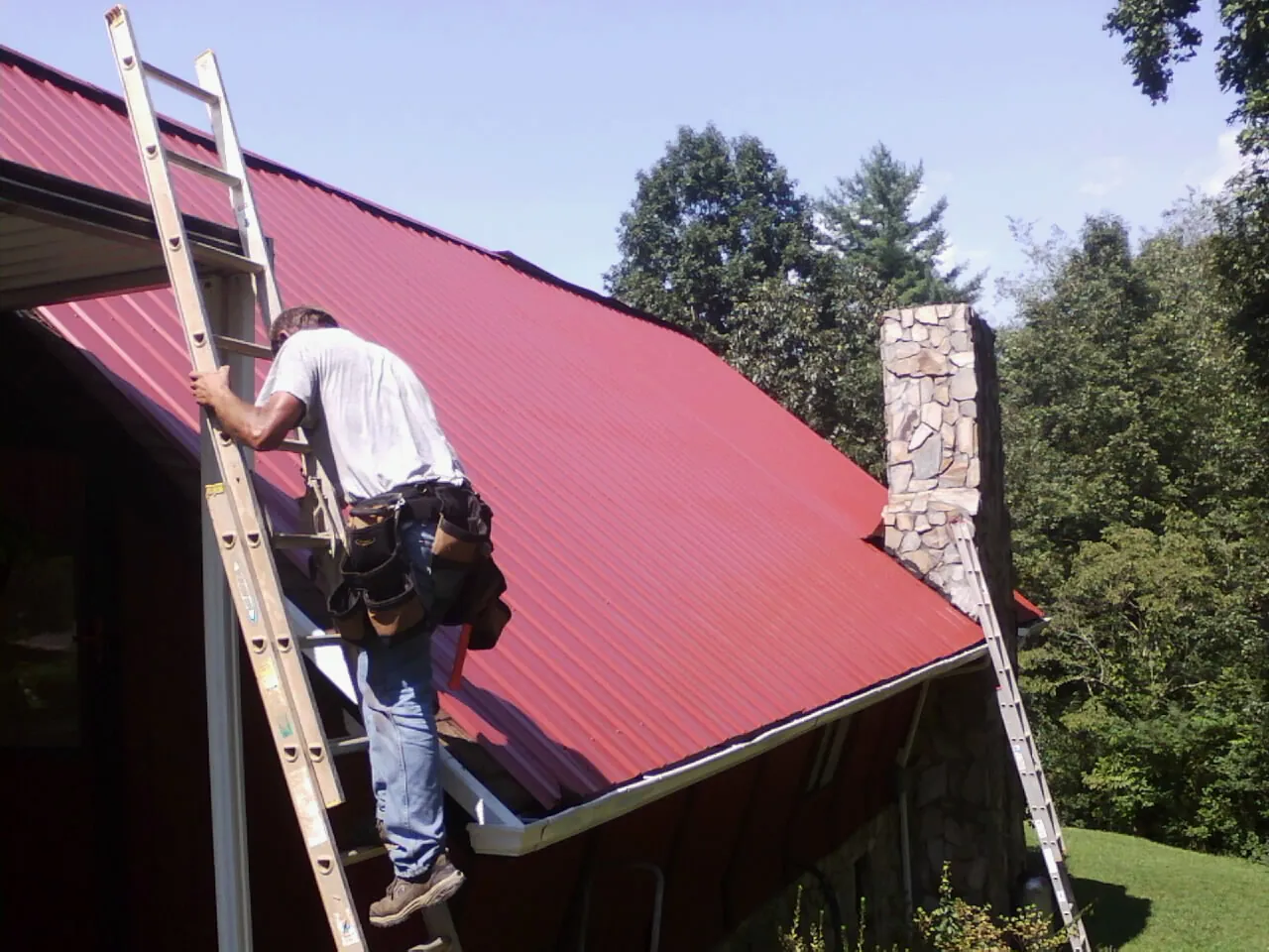 Roofer on a ladder beside a red metal roof and stone chimney — Soffit & Fascia Repair by Roofing Brothers in Griswold